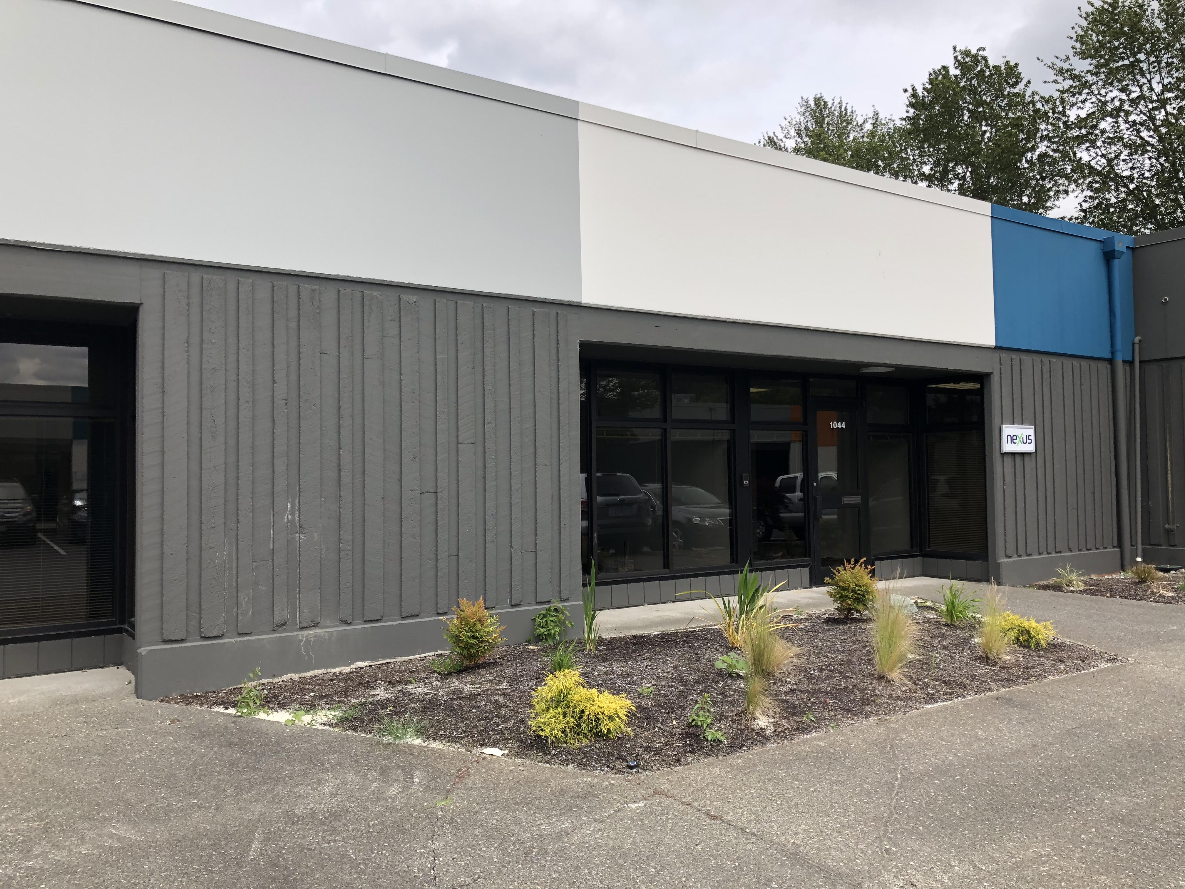 A single-story commercial building with gray and white exterior panels, large windows, and a small landscaped area with shrubs in front.